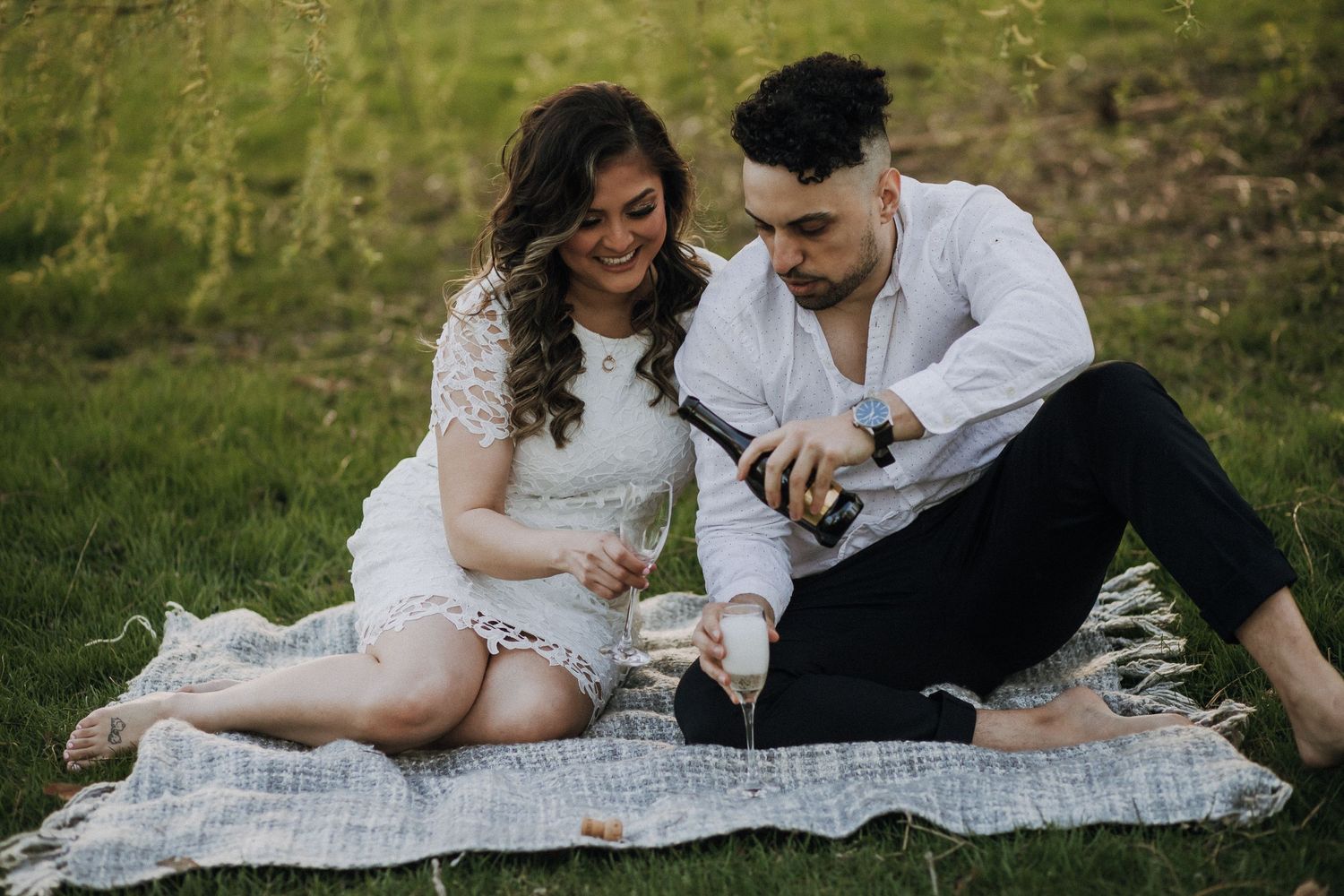 Couple having picnic on blanket in grassy field with drinks.