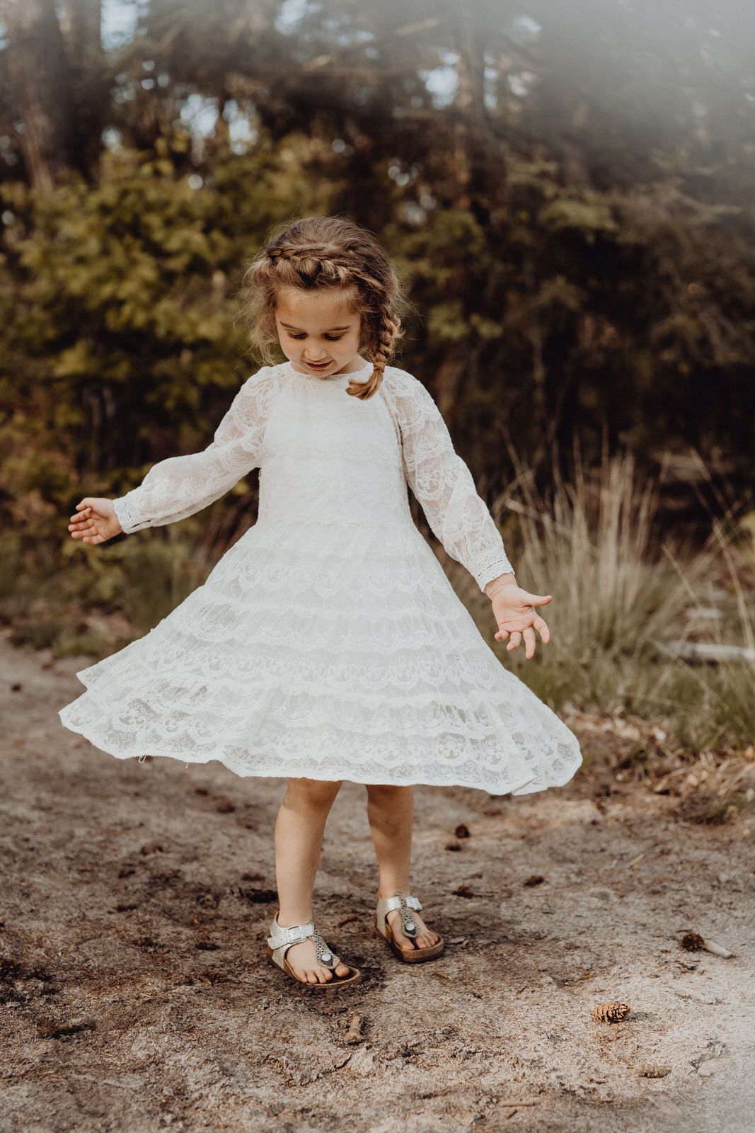 Child in white lace dress twirls joyfully on a dirt path surrounded by nature.