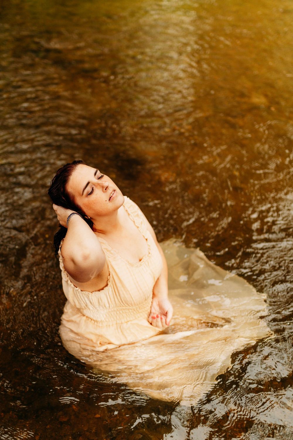 Young woman in cream dress reclines gracefully in shallow sunlit river with eyes closed.
