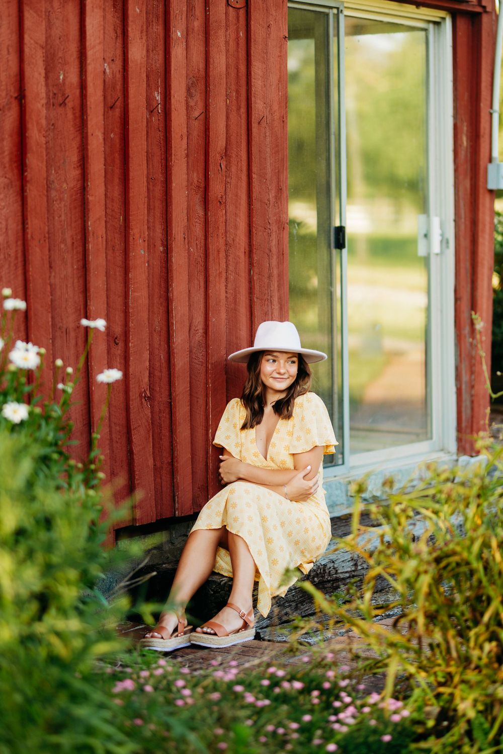 Smiling woman in yellow dress and white hat sits outside red wooden cabin with flowers.