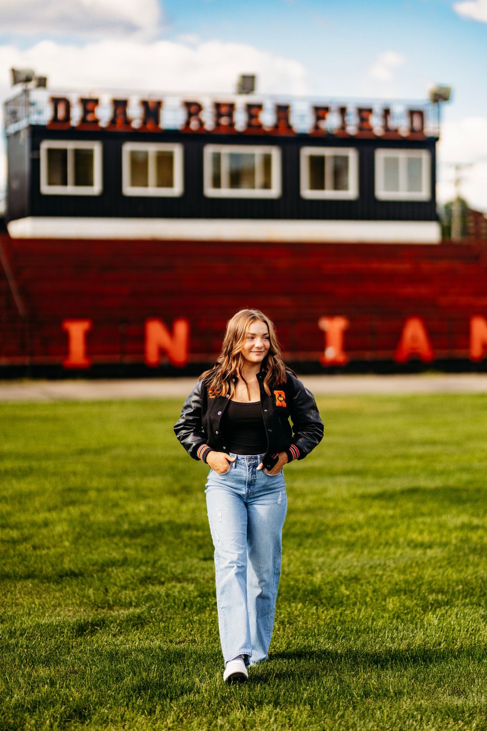 Teen girl in black varsity jacket and jeans walks across green football field at Dean Rhea Field.
