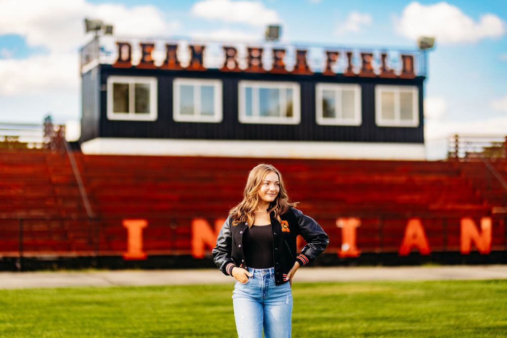 Senior girl smiling in black letterman jacket poses at Dean Rhea Field with Indians stadium backdrop.
