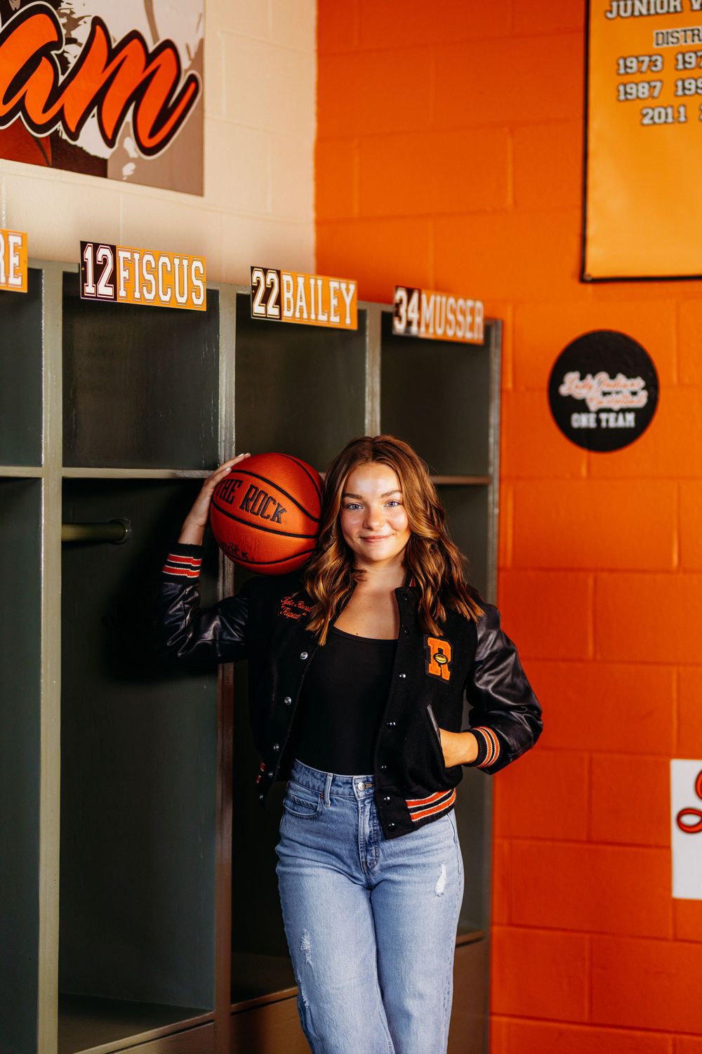 High school senior girl holding basketball poses by numbered lockers in orange and black locker room.