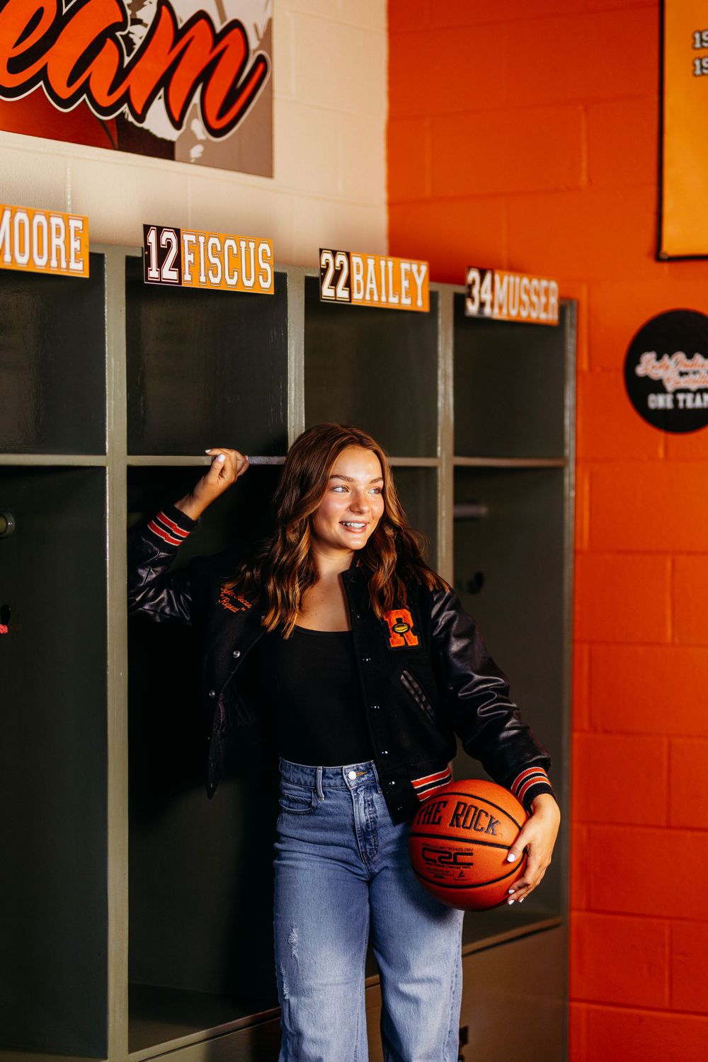 Smiling senior girl with basketball leans against team lockers in orange school locker room.