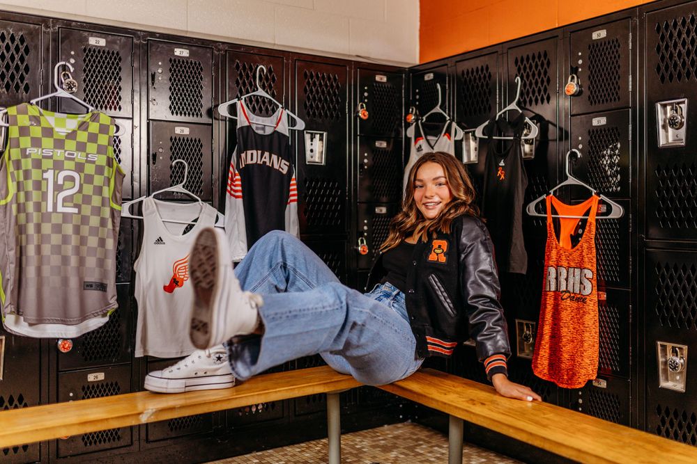 Senior girl in varsity jacket sits among basketball uniforms displayed in school locker room.