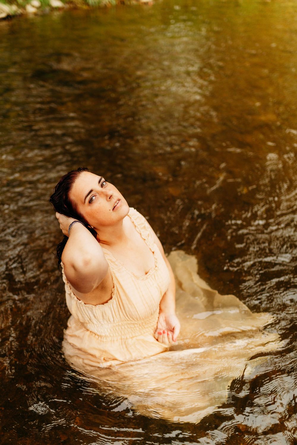 Young woman in wet cream dress sits in golden-lit river looking upward with serene expression.