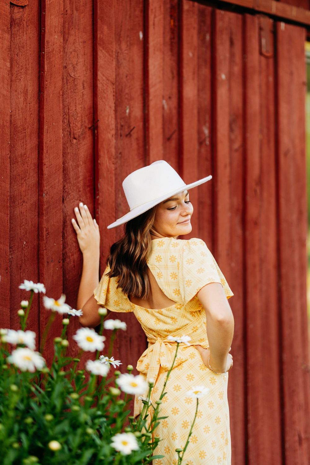 Woman in yellow polka dot dress and white hat poses by red wooden barn wall with daisies.