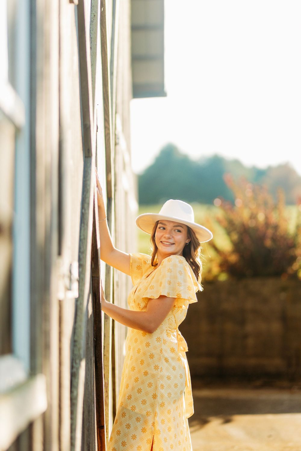 Young woman in yellow dress and white hat smiles while leaning on wooden barn in golden sunlight.