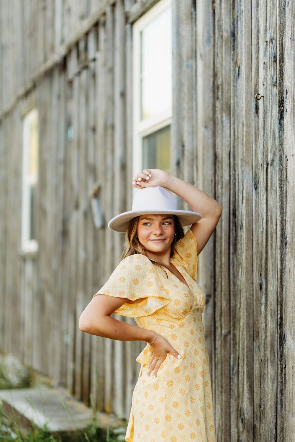 Smiling woman in yellow dress adjusts white hat while leaning against rustic wooden barn wall.