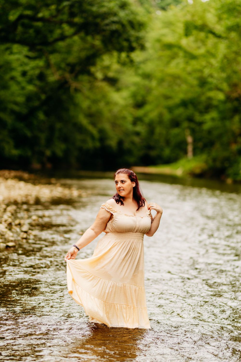 Young woman in flowing cream maxi dress wades through shallow river surrounded by green trees.