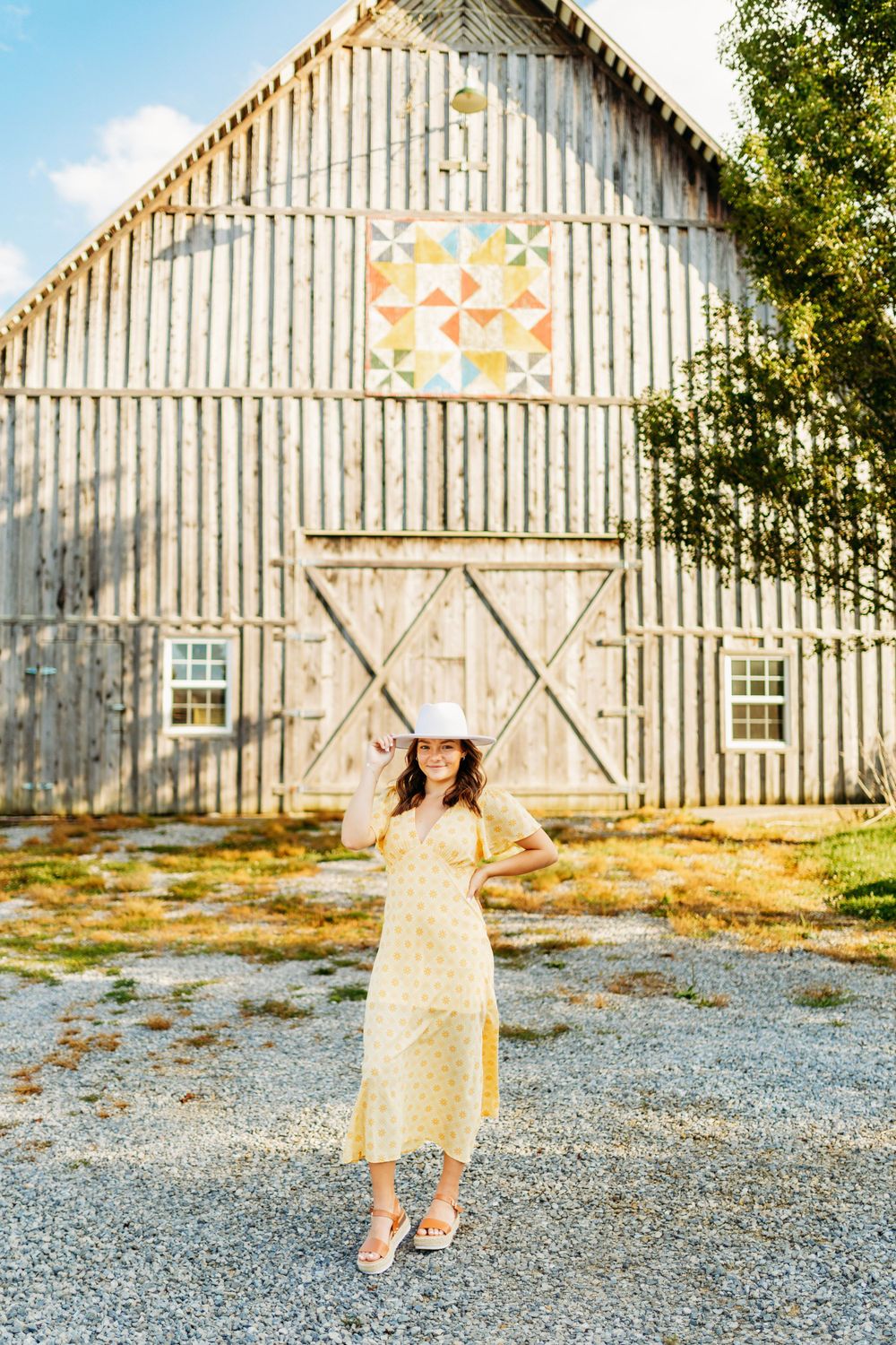 Young woman in yellow dress and white hat stands before large white barn with colorful quilt square.