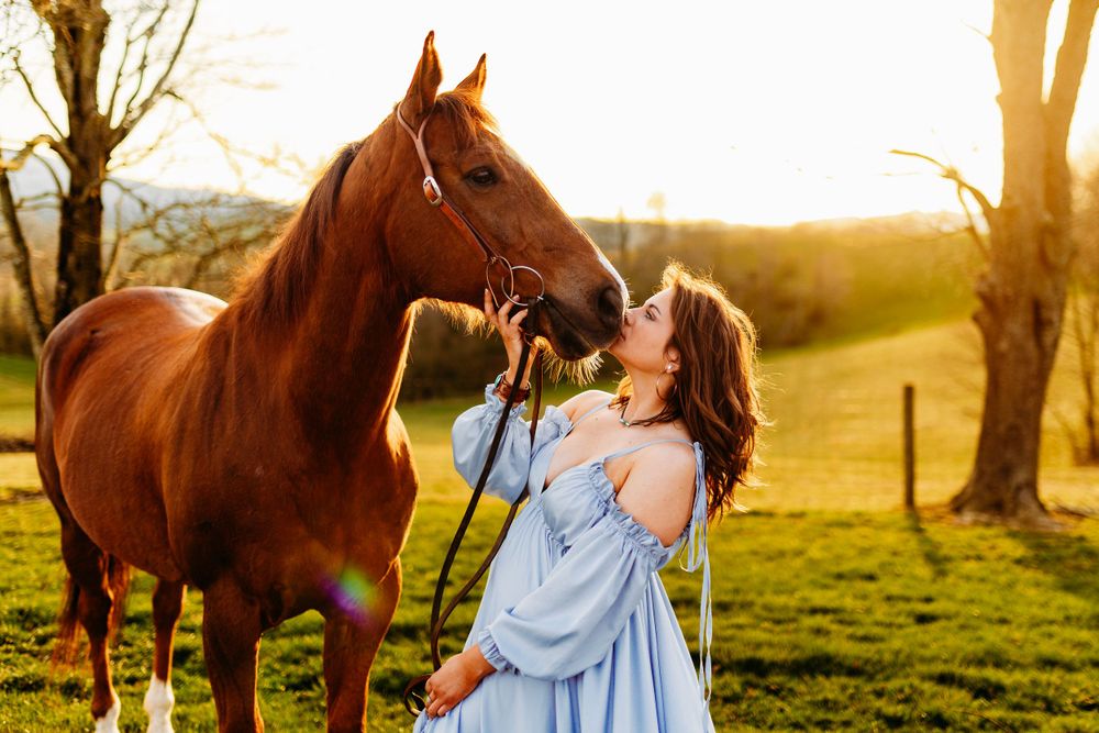 Woman in off-shoulder blue dress kissing chestnut horse at golden hour on green pasture.