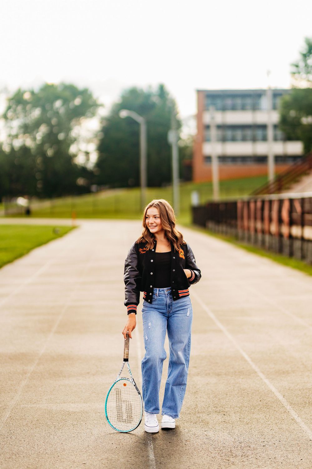 Teen girl in varsity jacket carries tennis racket while walking on outdoor track near school building.
