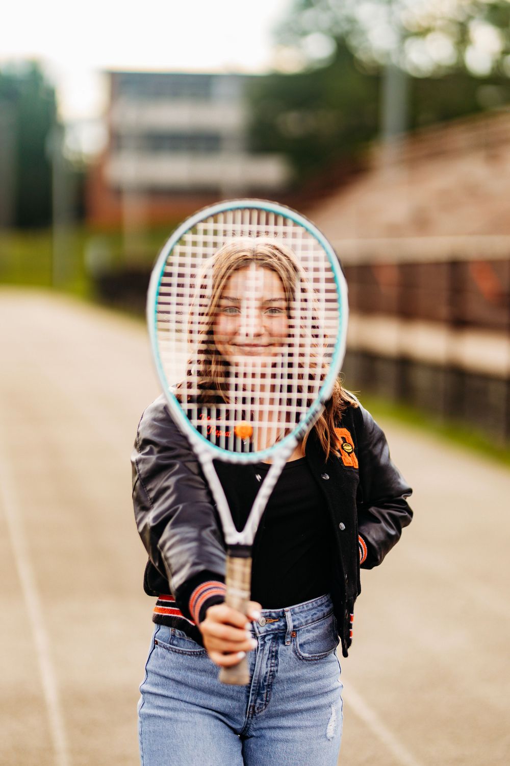 Smiling teen girl holds tennis racket up to her face on an outdoor court wearing a varsity jacket.