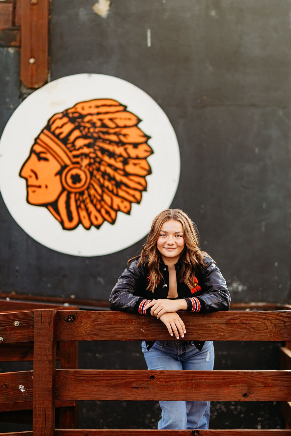 Teen girl in varsity jacket leans on wooden railing in front of Native American school mascot mural.