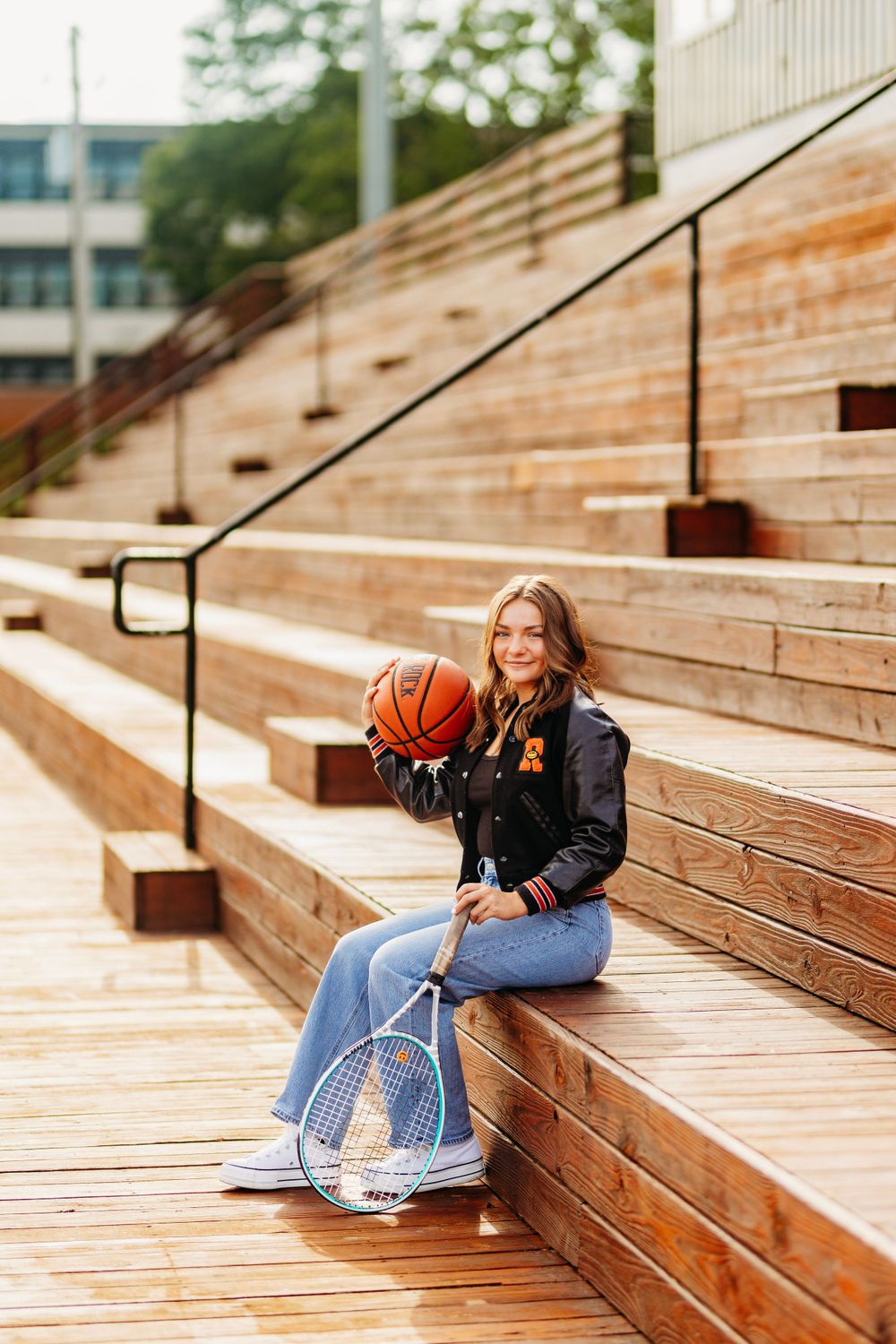 Teen girl in varsity jacket holds basketball and tennis racket while sitting on wooden bleachers.