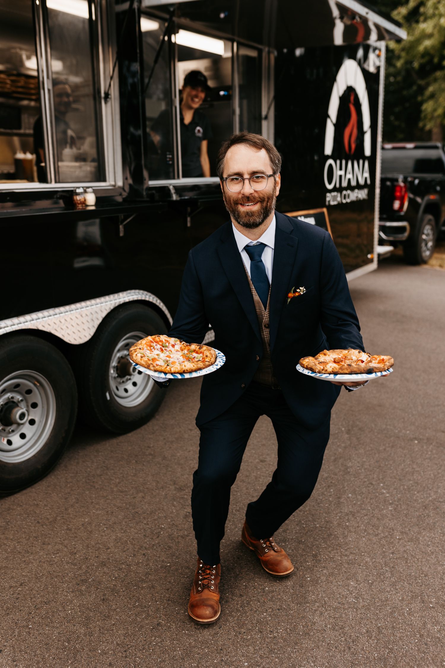 A man in a navy suit holding two pizzas in front of a food truck.