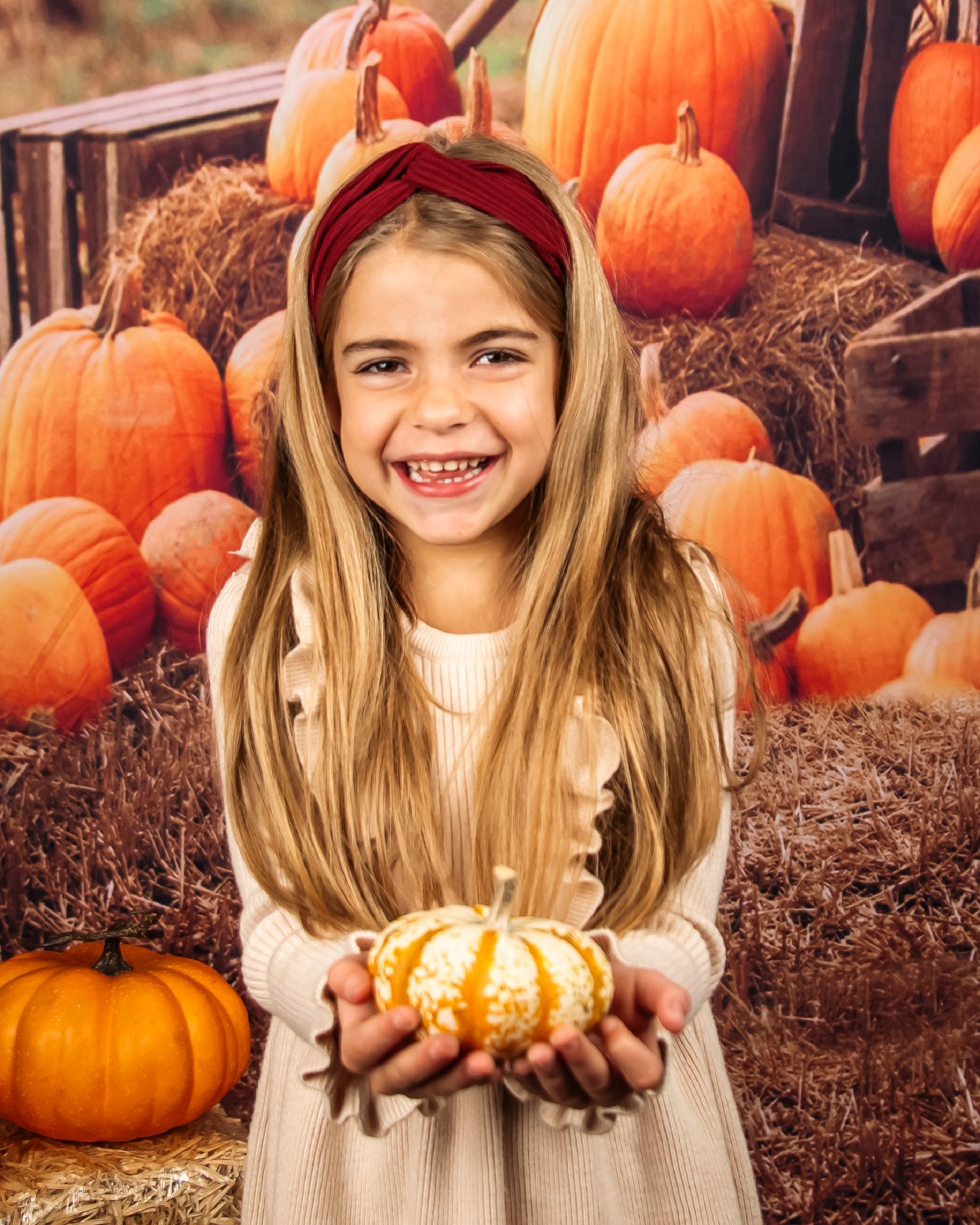 A child in a beige dress holds a small decorative pumpkin among larger orange pumpkins.