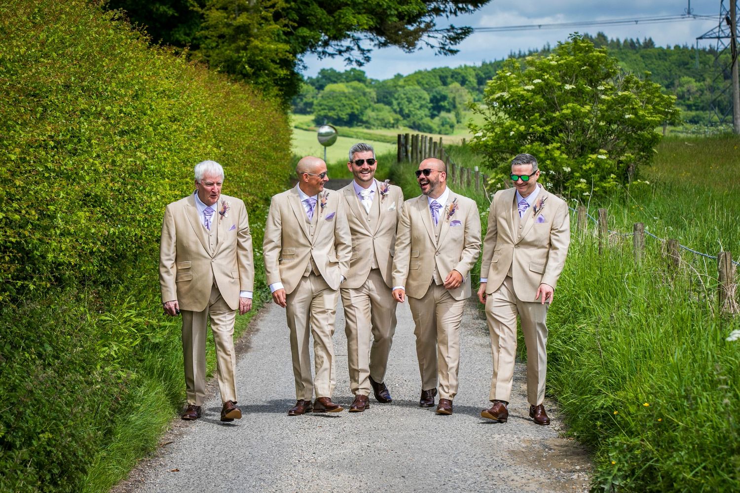 Groomsmen in tan suits pose together on a lush green hillside for wedding photos.