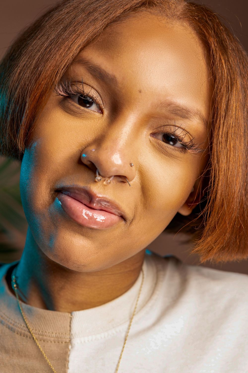 Close-up beauty portrait with natural makeup, nose ring, and auburn hair in soft lighting.