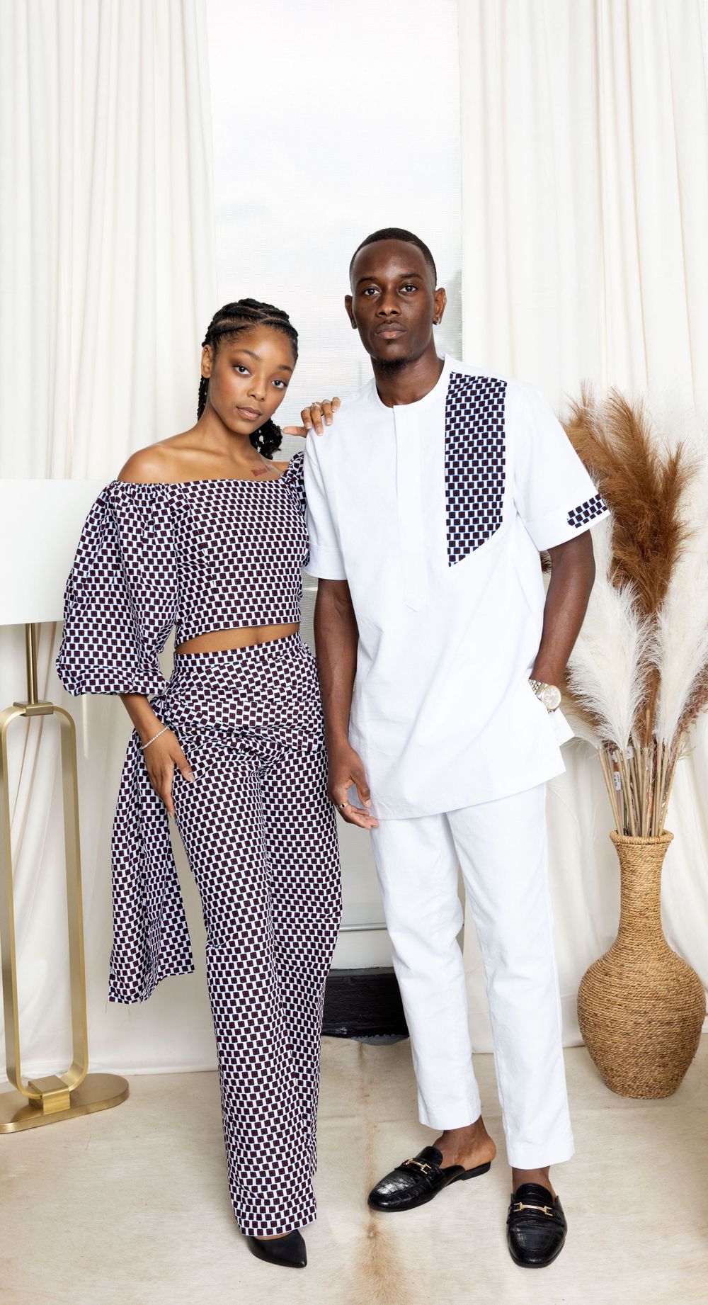 Couple in coordinated polka dot and white outfits pose against light curtain backdrop.