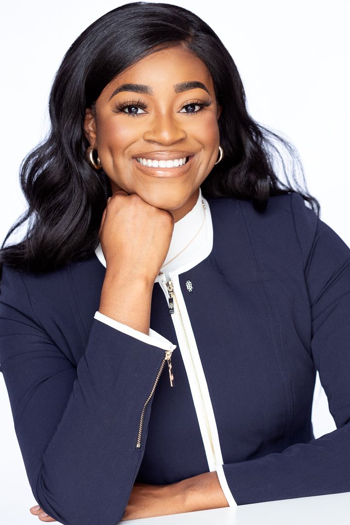 Professional headshot in a navy blazer with white trim showing a warm smile and wavy black hair.