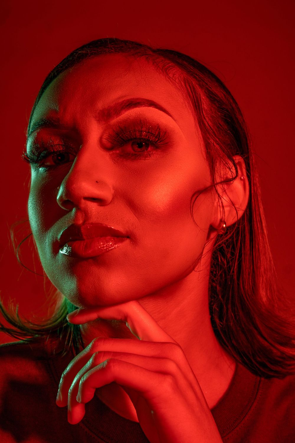 Dramatic portrait in red lighting with green accent showing artistic makeup and sleek hair.