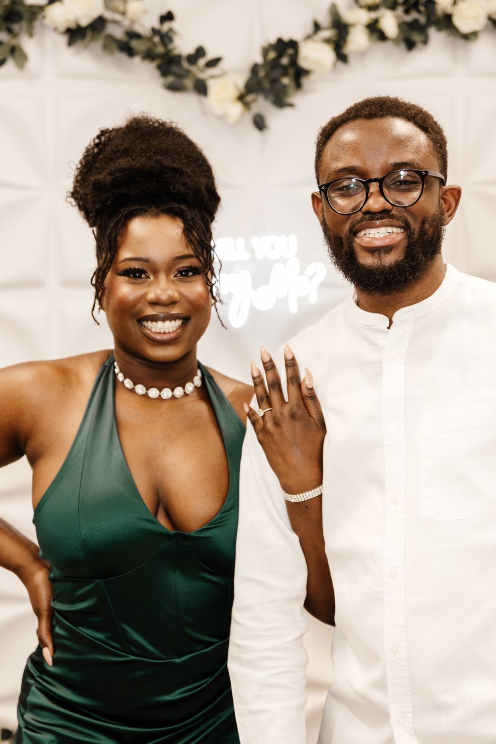 Engagement photo featuring couple in green dress and white outfit against floral backdrop.