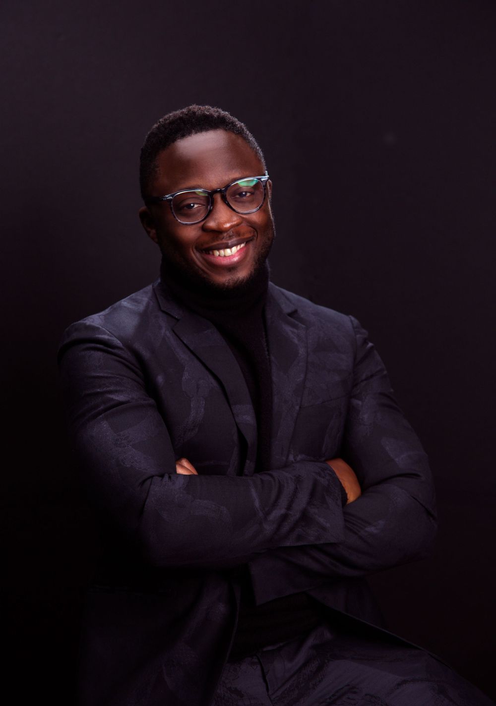 Professional portrait of a person in a black blazer and turtleneck smiling warmly against dark background.