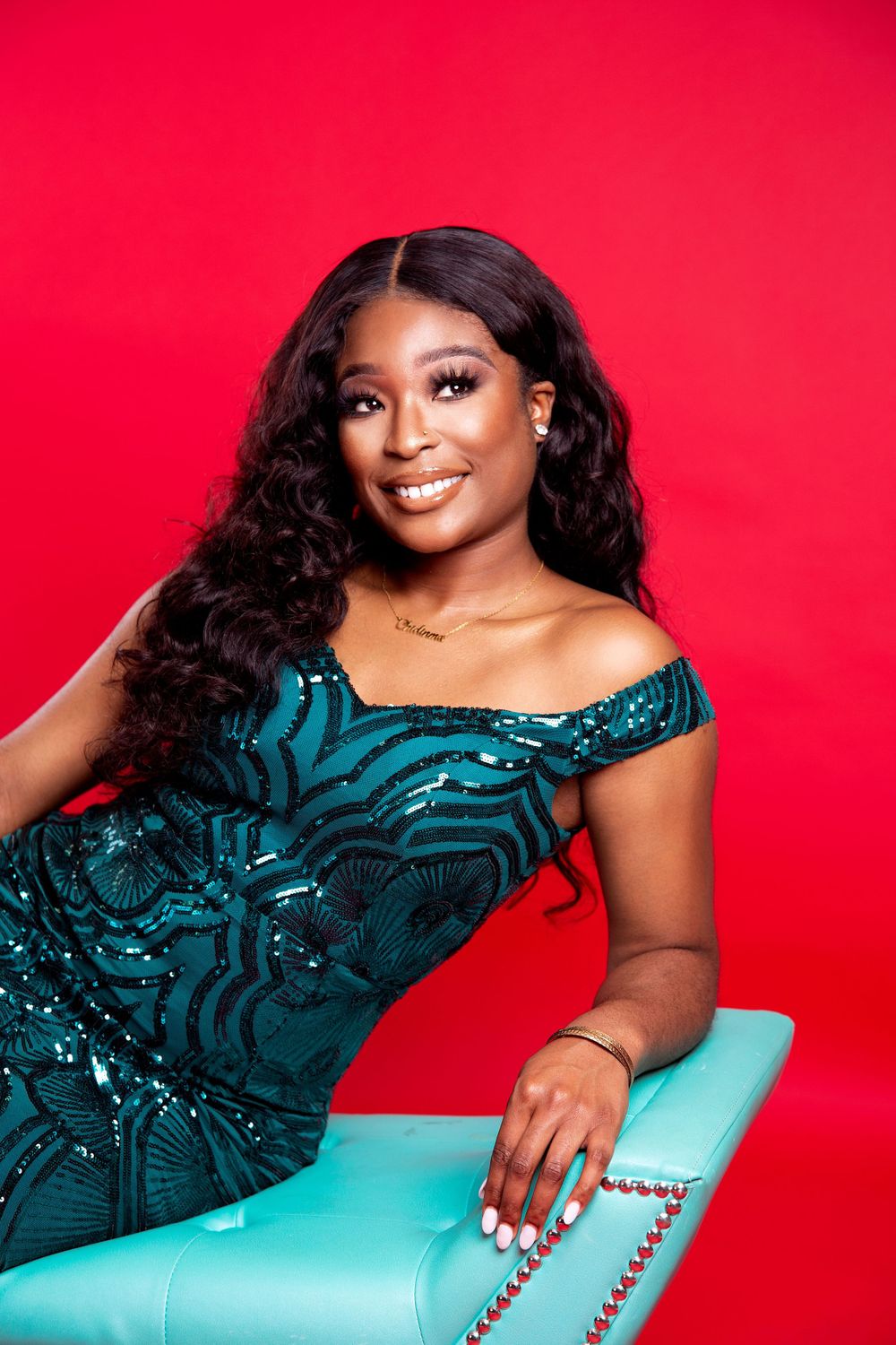 Woman in a teal sequined dress poses on turquoise furniture against a bright red background.