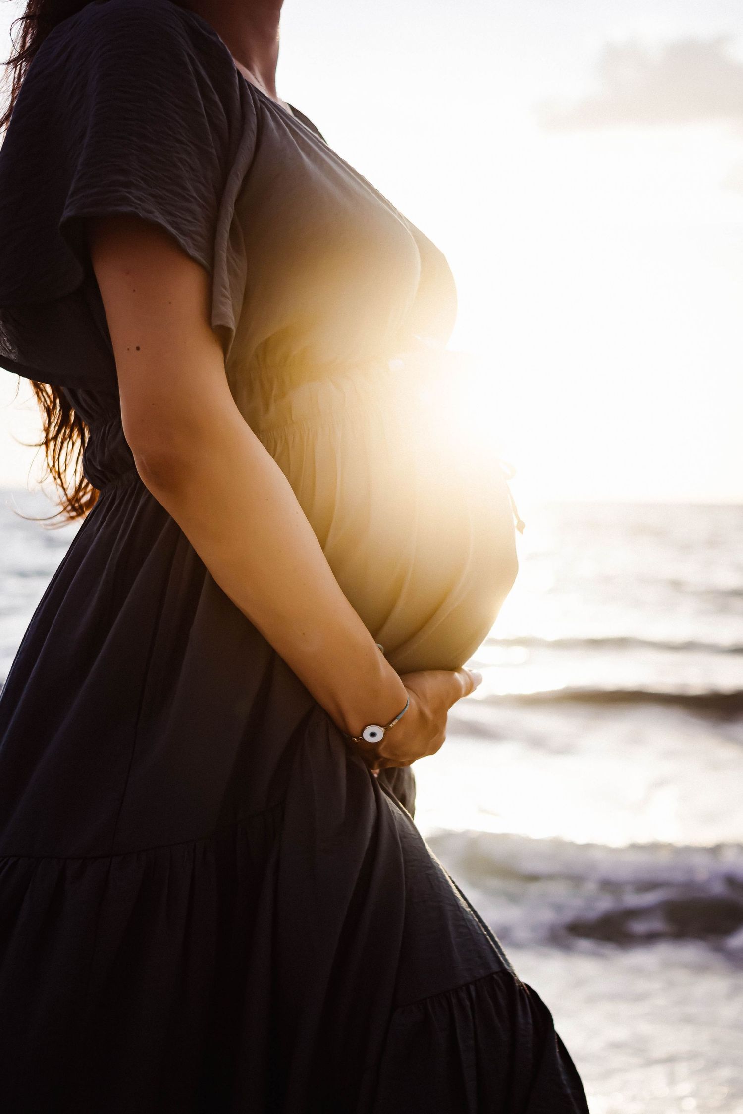 Silhouette of pregnant woman in black dress against ocean sunset.