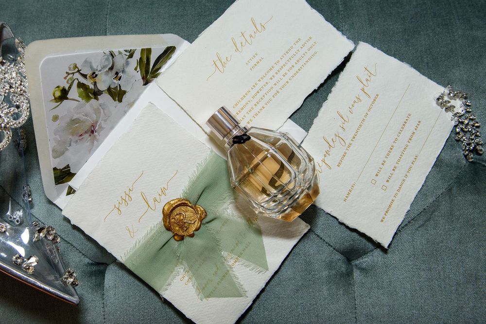 Elegant wedding invitation with perfume bottle and wax seal displayed on textured gray surface with white floral accents.