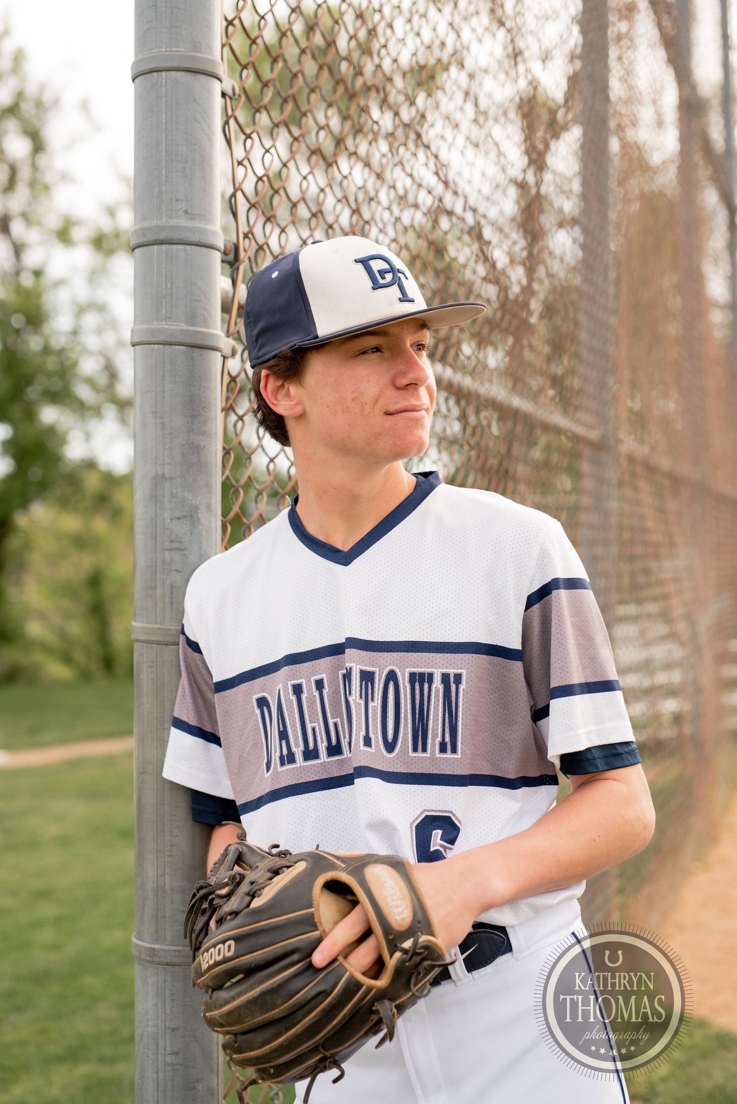 Baseball player in Dellasoft uniform poses with glove by chain-link fence.