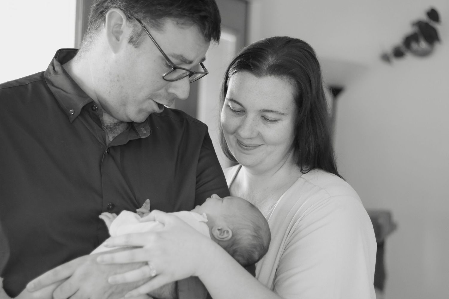 New parents, mum and dad holding newborn baby girl in their arms, smiling looking down at her.