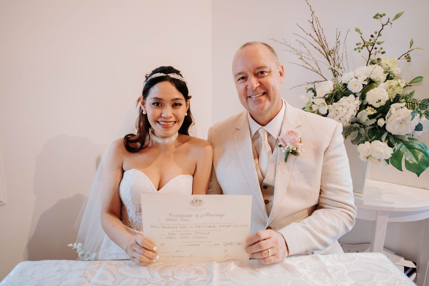 Newlyweds signing their marriage certificate together at a beautifully decorated ceremony table with floral arrangements.