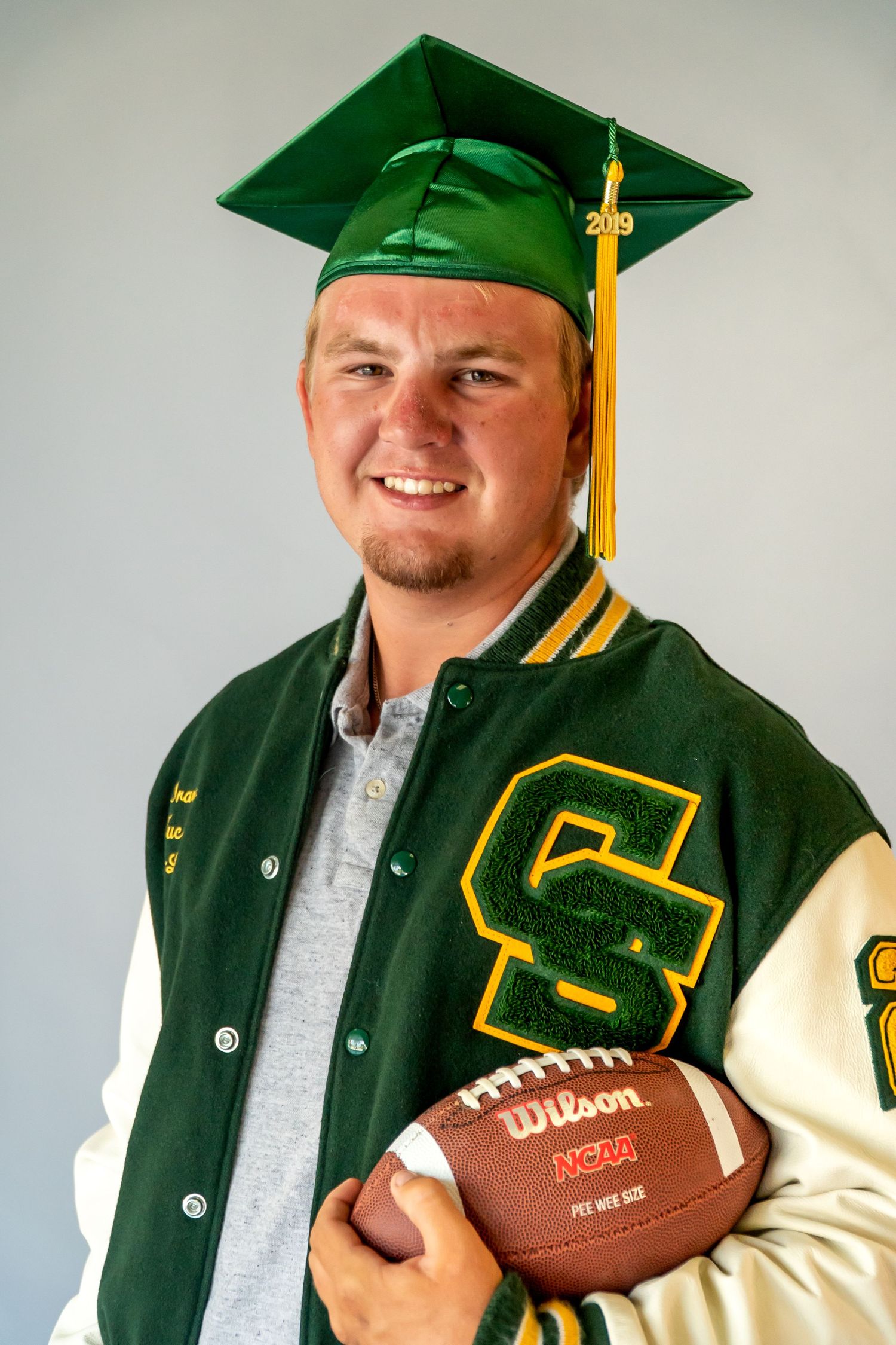 Graduate in green and gold academic regalia poses with a Wilson football showing school spirit.