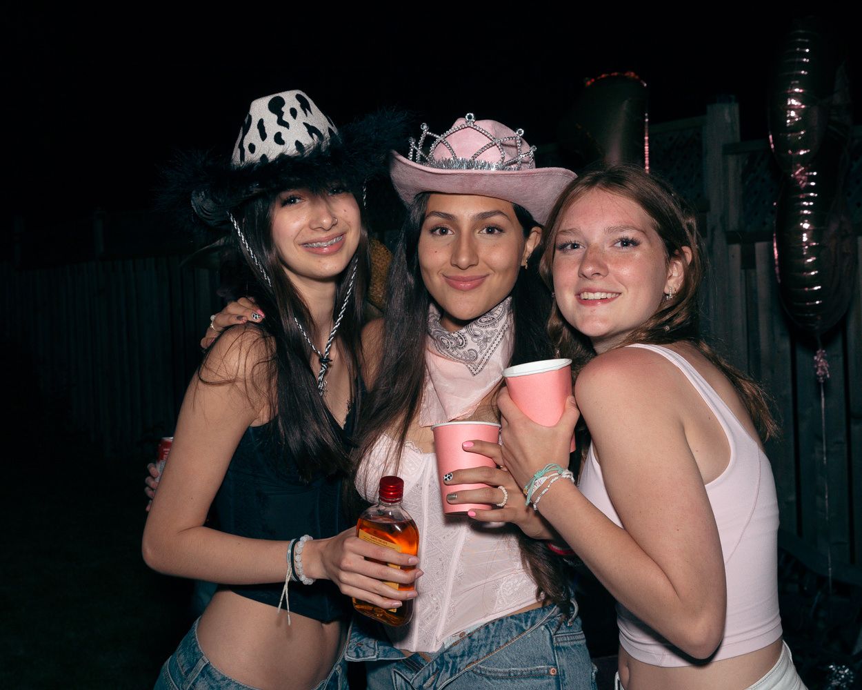Three friends in party attire and cowboy hats hold drinks while celebrating outdoors.