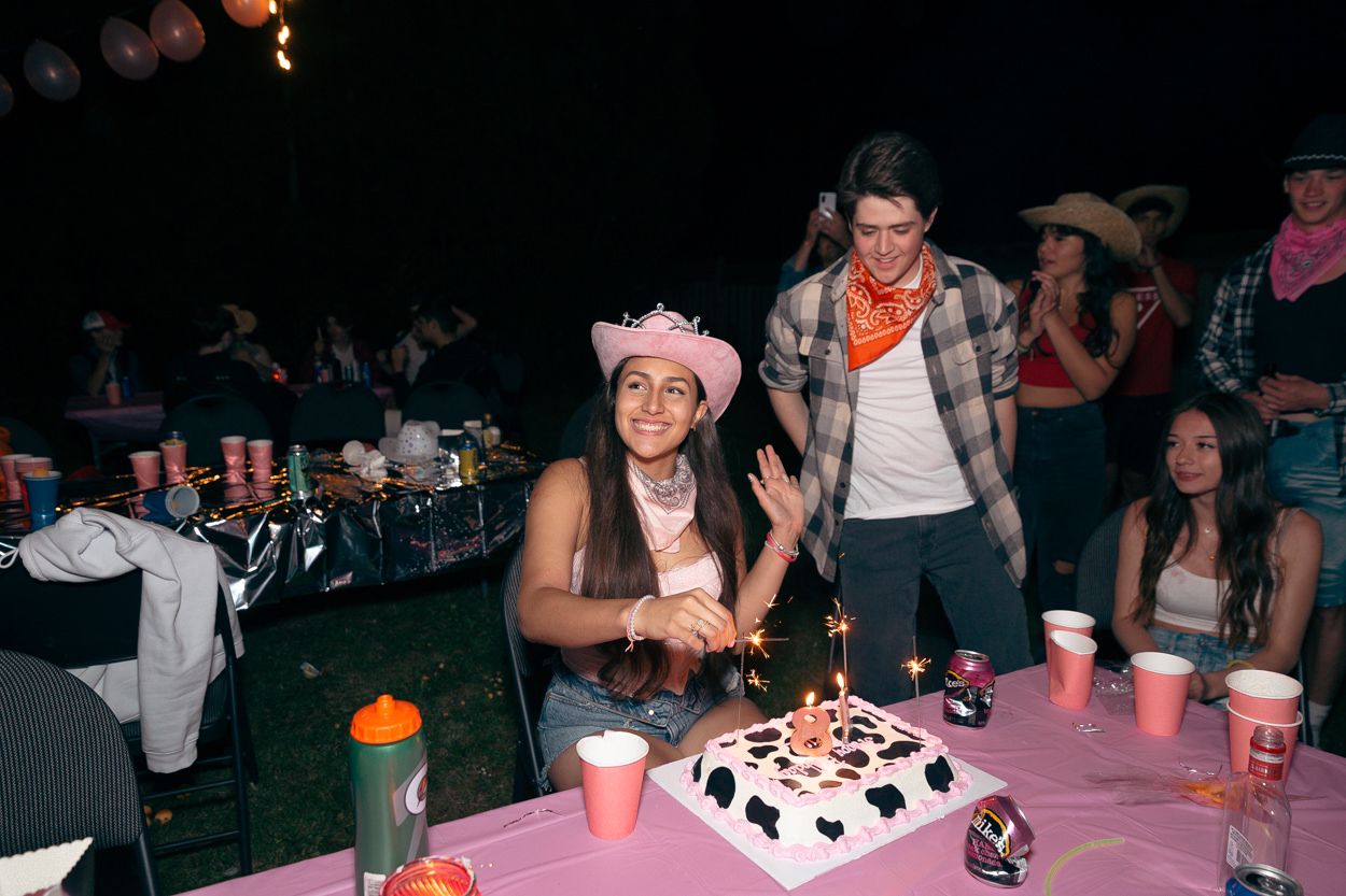 A birthday celebration at night with a lit cake on a pink table surrounded by party decorations.