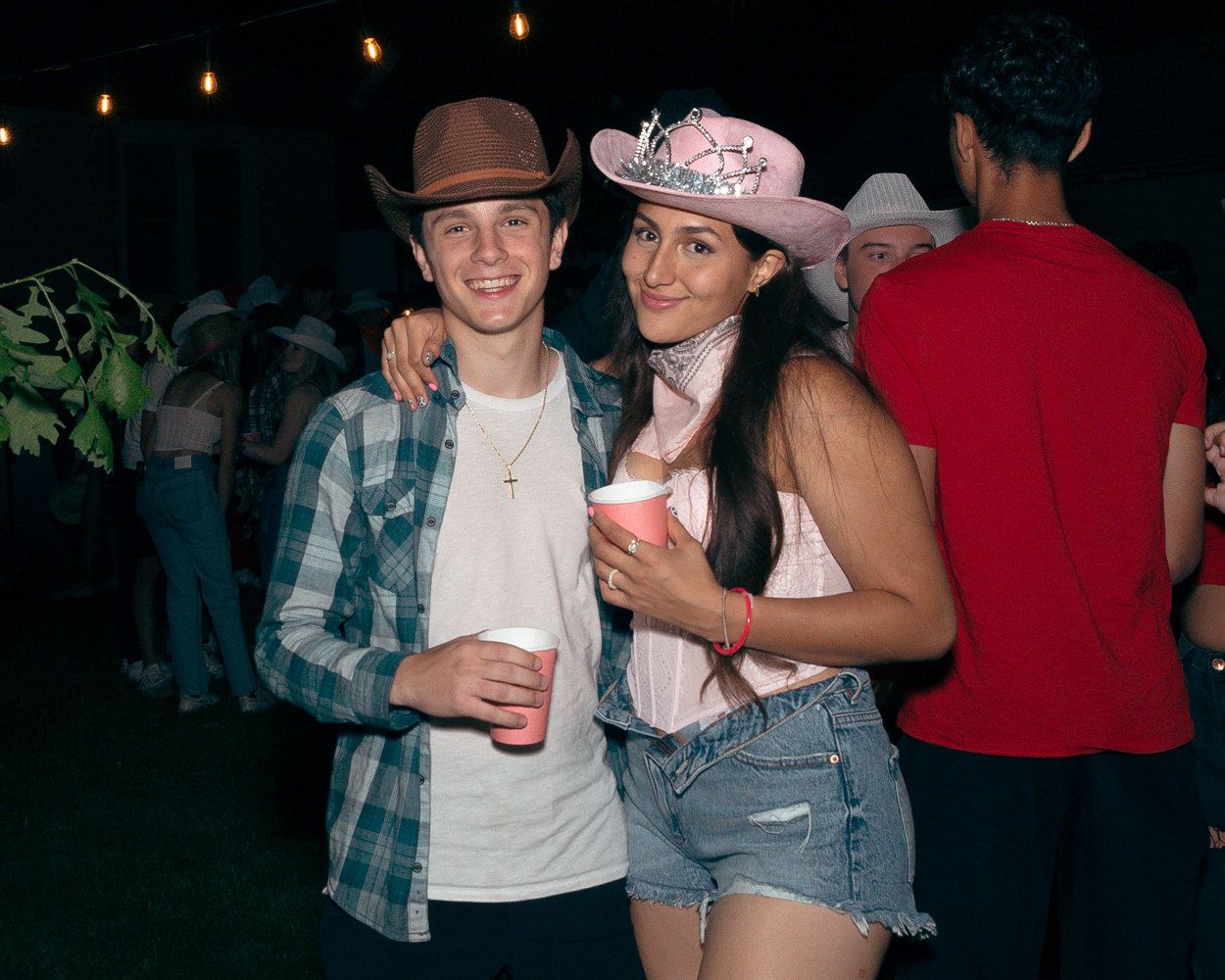 A couple poses at a western-themed party with string lights and foliage in the background.