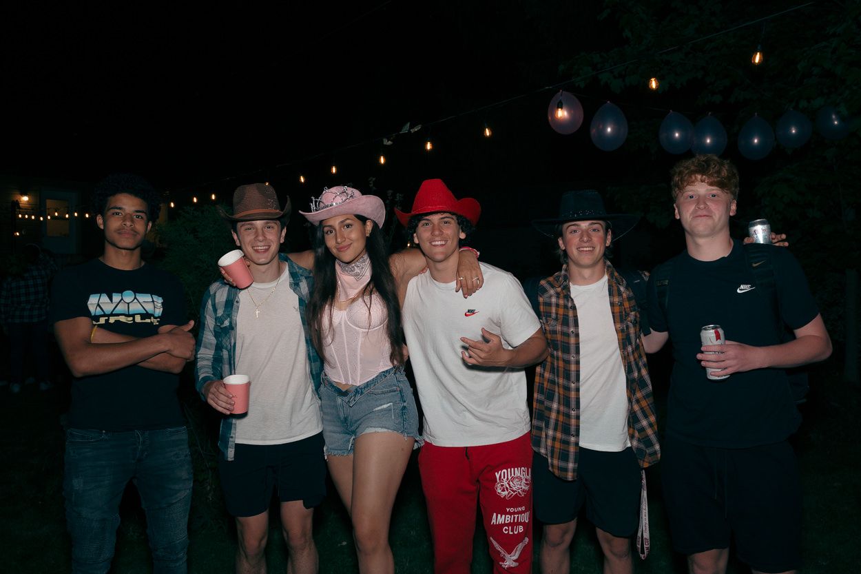 Group of friends at an outdoor party with string lights wearing casual western outfits and hats.