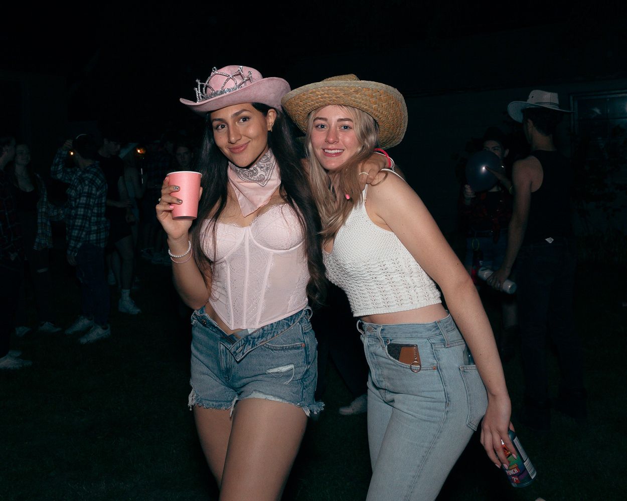 Two friends in western-inspired outfits hold drinks at an outdoor party with string lights.