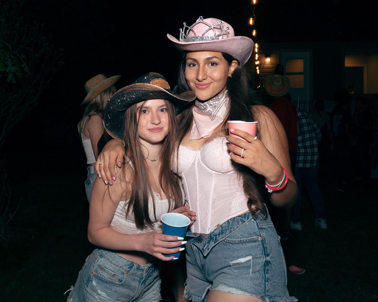 Two partygoers wearing cowboy hats and denim shorts pose together at a nighttime outdoor event.