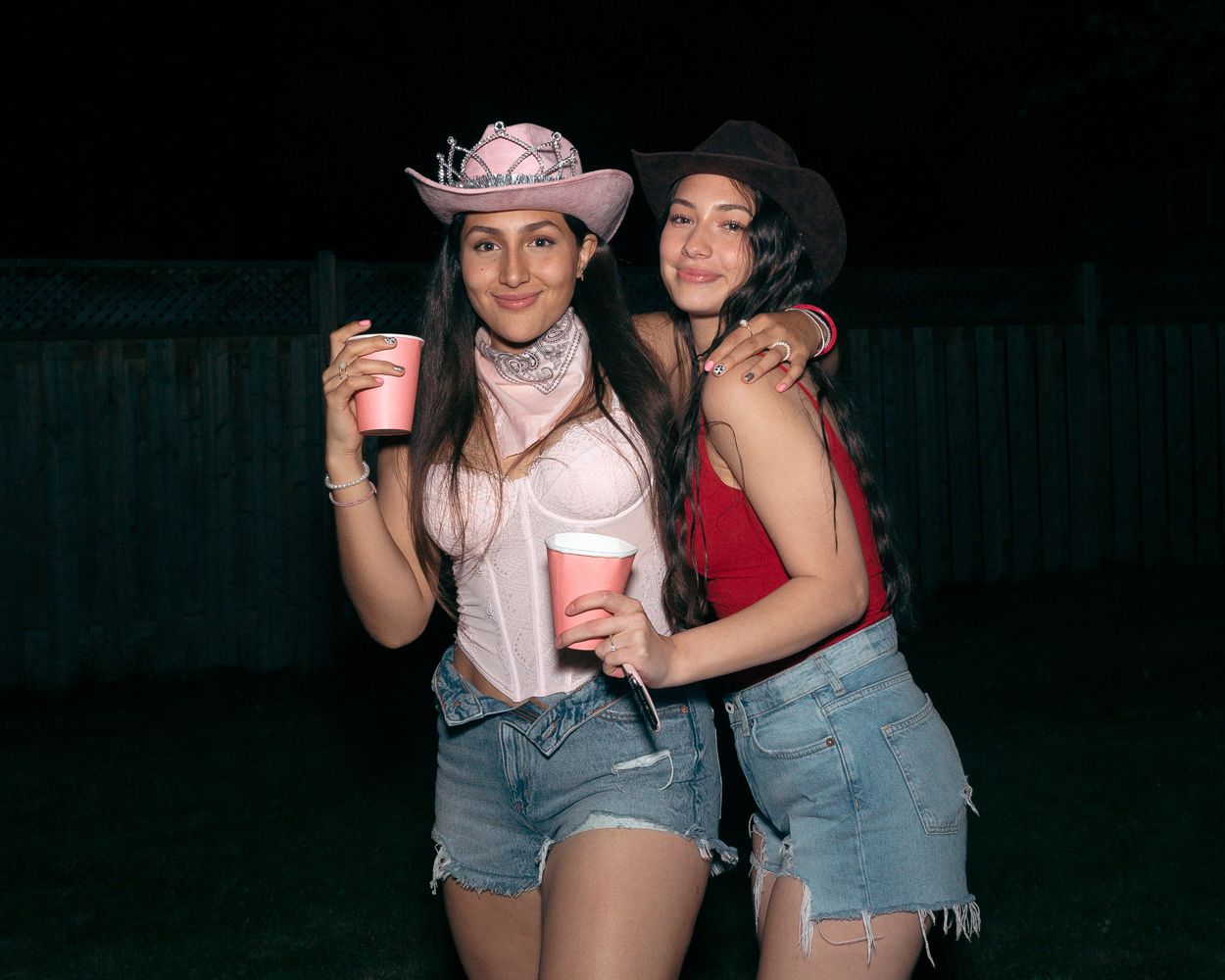 Two friends in casual western outfits pose with drinks at an outdoor party.