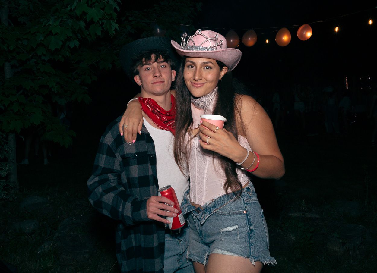 Two friends in western attire pose together at night with drinks and string lights in the background.