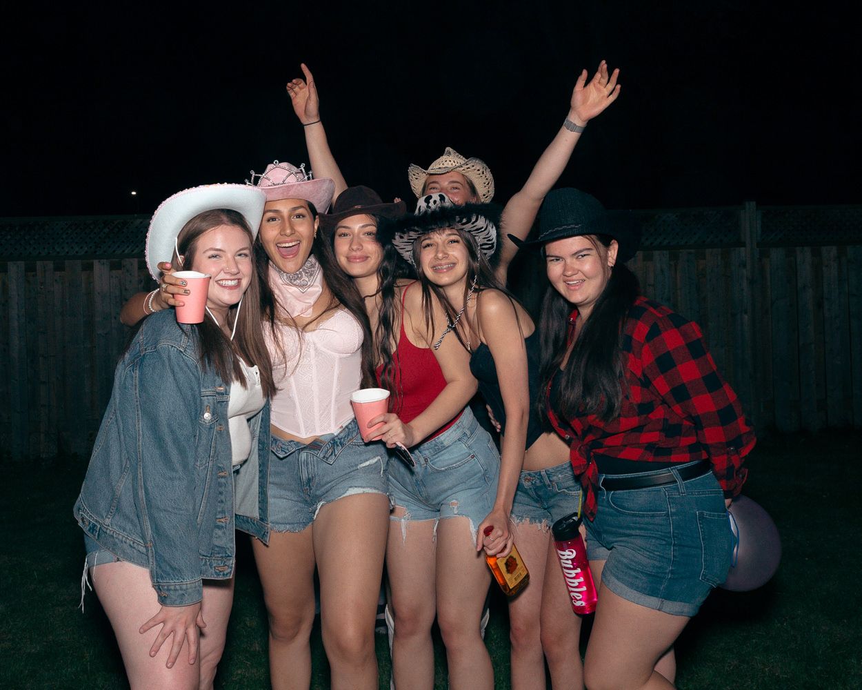 Friends in western-themed outfits celebrating at a backyard party with string lights.