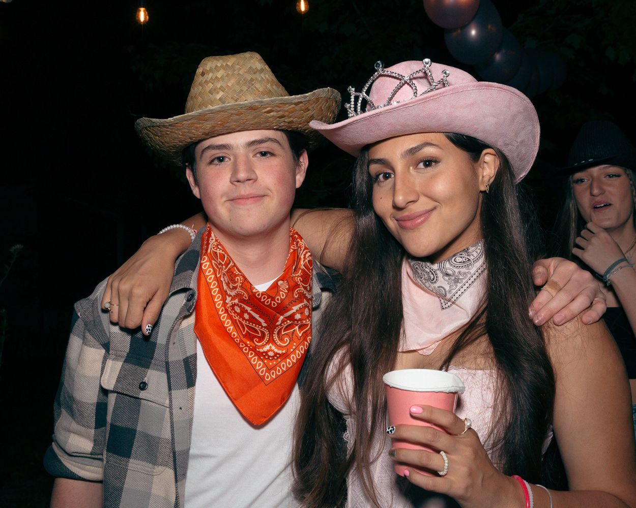 Party-goers in western attire including bandanas and cowboy hats pose together at night.