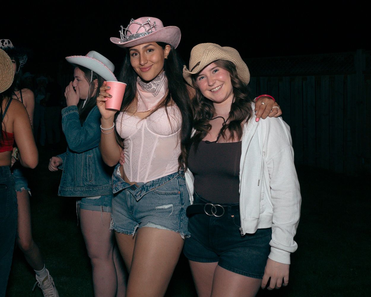 Two friends in western-inspired outfits pose with drinks at a nighttime outdoor party.