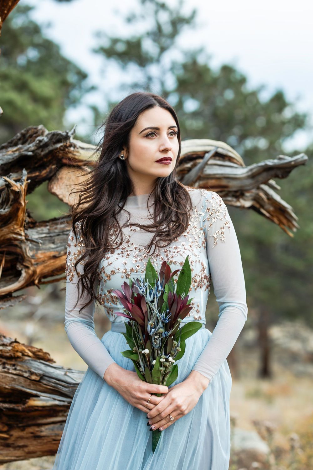 A woman in a pale blue dress holds native flowers against rustic wooden scenery.