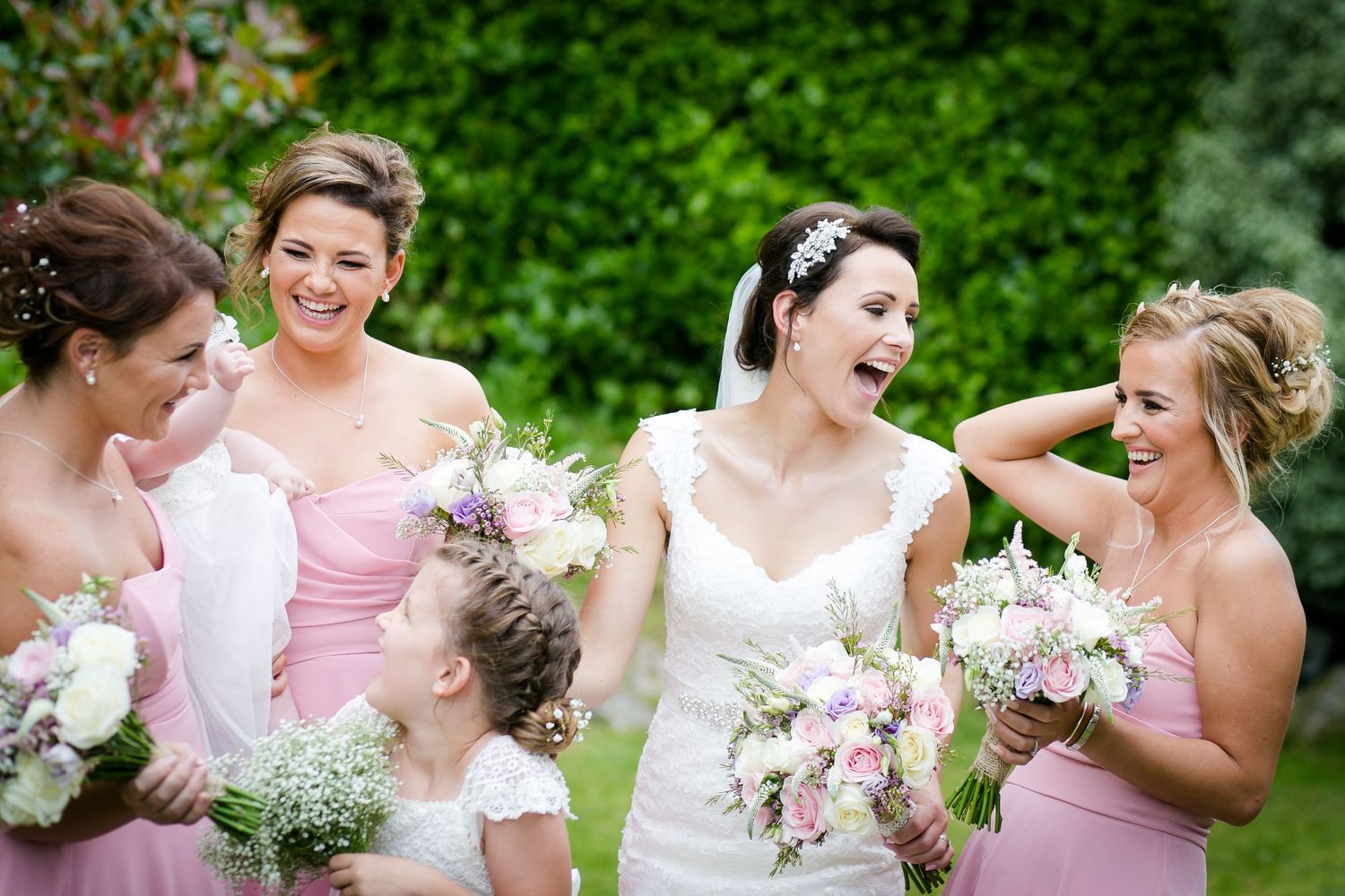 Bride laughing joyfully with bridesmaids in pink dresses holding bouquets outdoors.