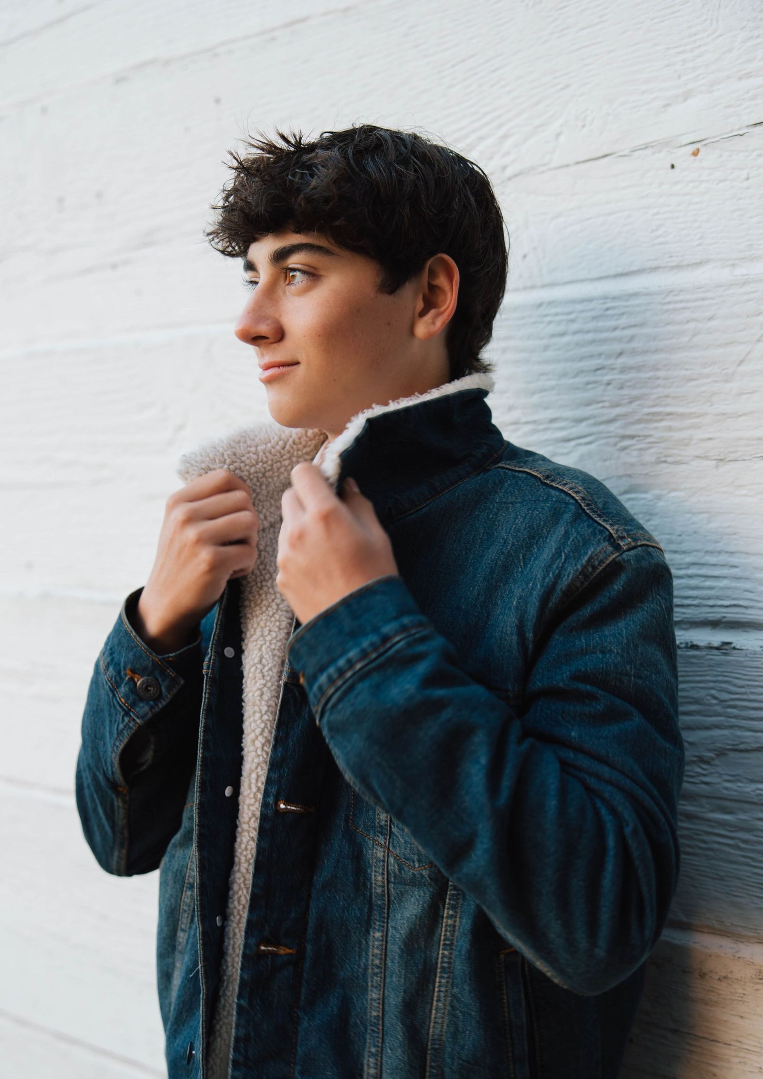 Teen boy in denim sherpa jacket adjusts collar while leaning against white brick wall.